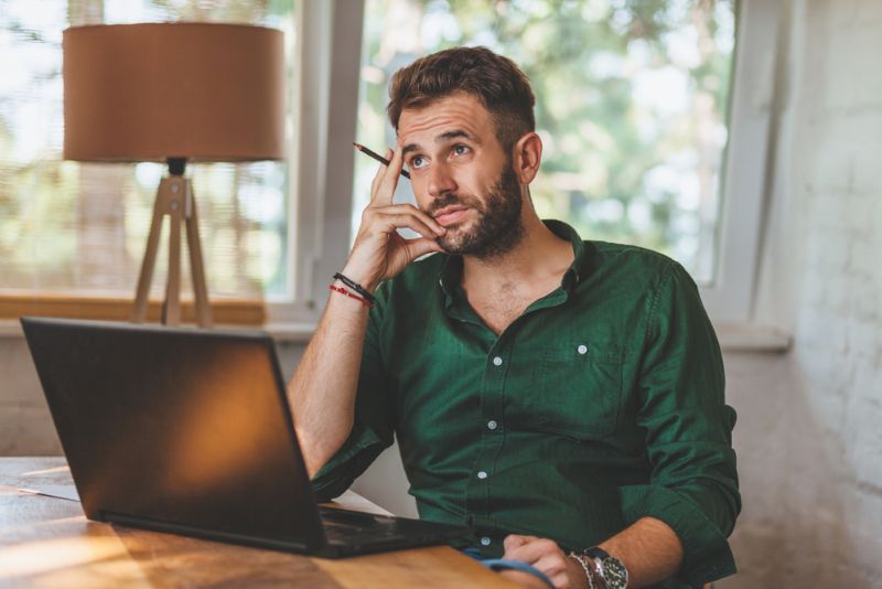 Young man sitting pensively at laptop with pencil in hand